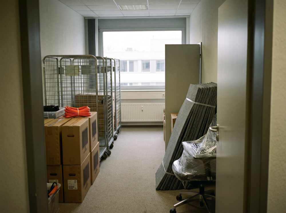 Büroraum vor der Verlagerung: Rollcontainer, beschriftete Kartons, gestapelte Teppichfliesen und ein in Folie gewickelter Bürostuhl.